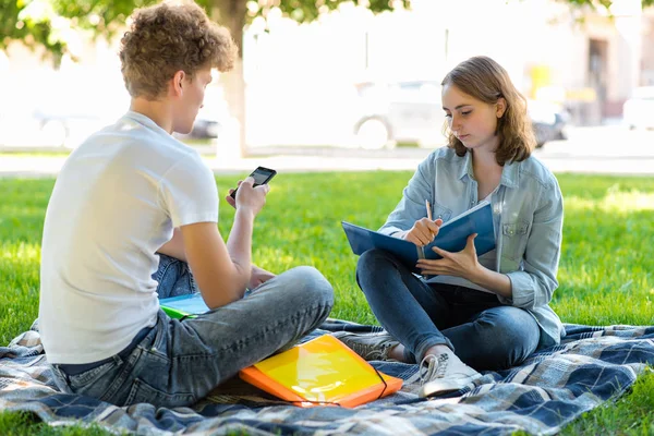 Ein Kerl mit einem Mädchen im Sommer in der Natur. Sie sitzen an der frischen Luft. Unterricht nach der Schule. Hände, die ein Smartphone halten, eine große Zusammenfassung. Hausaufgaben erledigt. — Stockfoto