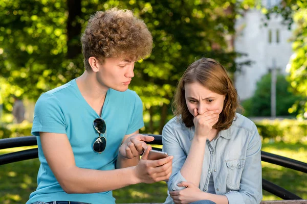 Un ragazzo con una ragazza in estate all'aperto. Un ragazzo si tiene per mano con uno smartphone che punta il dito contro il telefono. Emozioni di sfiducia. Il problema è in relazione. Rettifiche nella corrispondenza . — Foto Stock