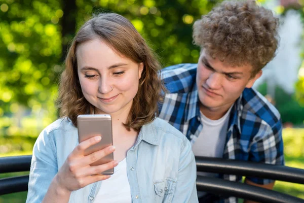 Una ragazza legge messaggi al telefono. Il tizio si sta spiando le spalle. Estate nella natura. Il concetto di sfiducia spiare l'interlocutore . — Foto Stock