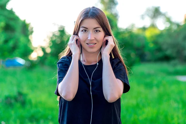 Vacker brunett flicka på sommaren i en park utomhus i en svart T-shirt. Ler tittar på ram på tänderna av tandställningen. Hon sätter på hörlurar och lyssnar på musik. — Stockfoto