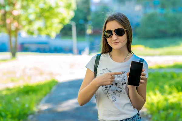 Een tienermeisje in haar armen heeft een smartphone. In de zomer in het park. Zonnebril. Vrije ruimte voor tekst. Concept van een nieuwe telefoon, applicatie op de telefoon. — Stockfoto