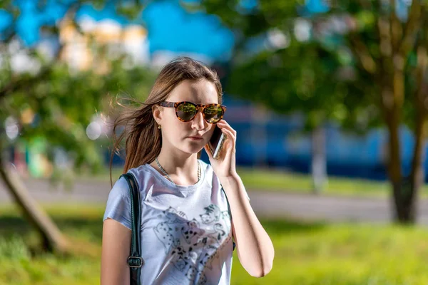 Una adolescente. Verano en la naturaleza. En sus manos tiene un smartphone. Habla por teléfono. Un día soleado en la ciudad. Vestido con una camiseta y gafas de sol . — Foto de Stock