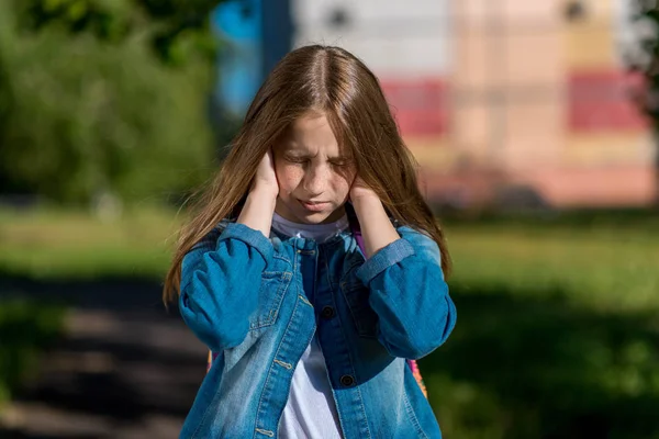 Little girl schoolgirl in summer park. He covers his ears with hands. The concept of loud noise, problems in family, scandal, reluctance listen, discontent. Emotions of quarrel, frustration, conflict. — Stock Photo, Image