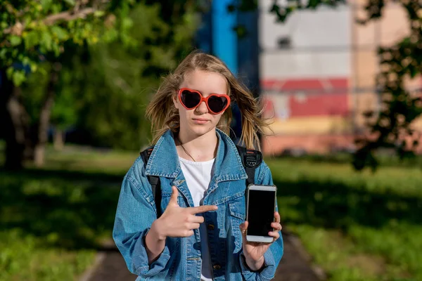 Dívka dospívající školačka. Léto v přírodě. Sluneční brýle ve tvaru srdce. Ve svých rukou drží smartphone. Koncept nového telefonu, nové aplikace. Volné místo pro text. — Stock fotografie