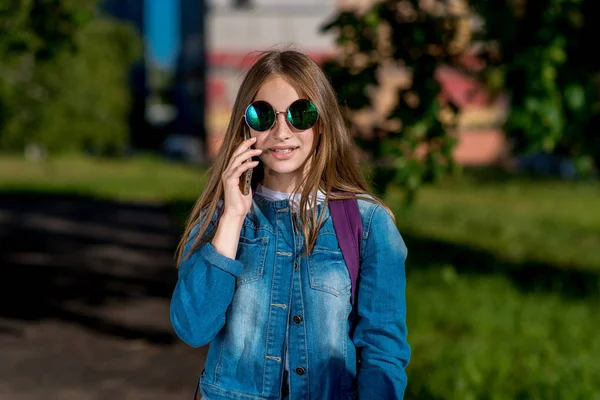 Gadis sekolahan remaja. Di musim panas di kota. Di tangannya memegang smartphone. Dalam pakaian jins, kacamata hitam. Dia berbicara di telepon, mendengarkan pesan di mesin penjawab . — Stok Foto