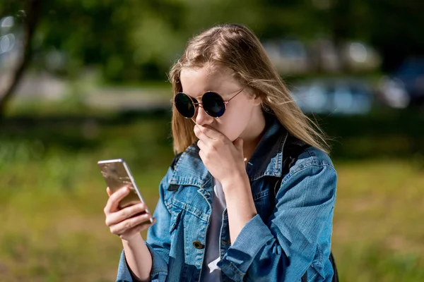 Gadis kecil remaja. Di musim panas di taman. Di tangannya memegang smartphone, dengan jaket denim dan kacamata hitam. Konsep emosi, kejutan, ketakutan, frustrasi, kecemasan, masalah . — Stok Foto