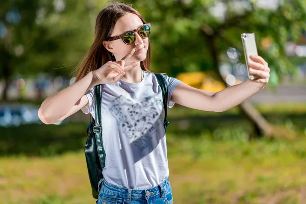 Nastolatek uczennica dziewczyna w parku lato za plecak, Okulary przeciwsłoneczne. W jego rękach posiada smartfona. Koncepcja wideorozmowy na telefon, robić zdjęcia o sobie na smartfonie. Uśmiecha się radośnie. — Zdjęcie stockowe