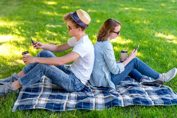 Young student girl and boy. They are sitting in park in nature. In the summer on a plaid. In hands of smart phones. Watching video on the phone. Camping. In hands of coffee mugs with hot coffee.