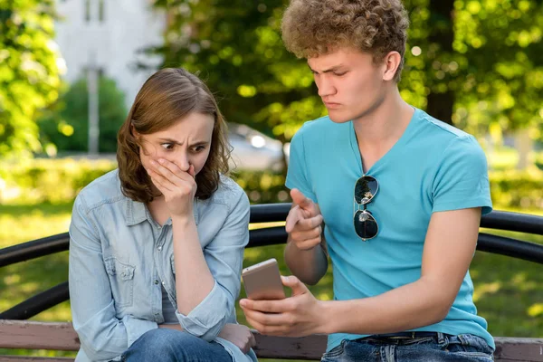 A young man points a finger at a smartphone girl. The problem is in the relationship. Emotions of mistrust. Concepts of betrayal and betrayal. Young couple in summer in a park outdoors. — Stock Photo, Image