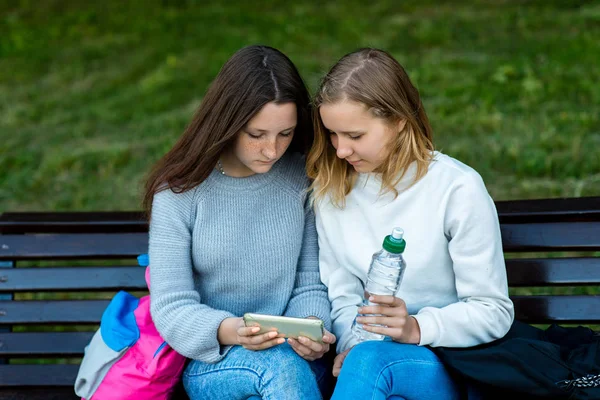 Twee schoolmeisjes student. In de zomer zitten ze op een bankje. Zij rusten na school in de natuur. Handen met een fles water. Kijken naar video op telefoon. — Stockfoto