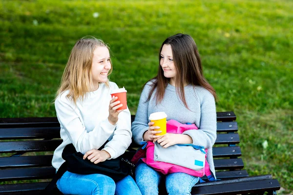 Две девочки школьницы. Летом сидя на скамейке природы. Он держит в руках горячий кофе или чай. Счастливо общается после уроков. Понятие "лучшие друзья" - это эмоции удовольствия от счастья . — стоковое фото