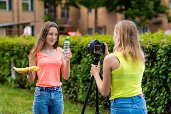 Deux petites amies, l'été en ville. Enregistrez la vidéo sur la caméra. Dans les mains tenant une bouteille d'eau de banane. Le concept de saine alimentation. Utiliser un appareil photo avec trépied . — Photo