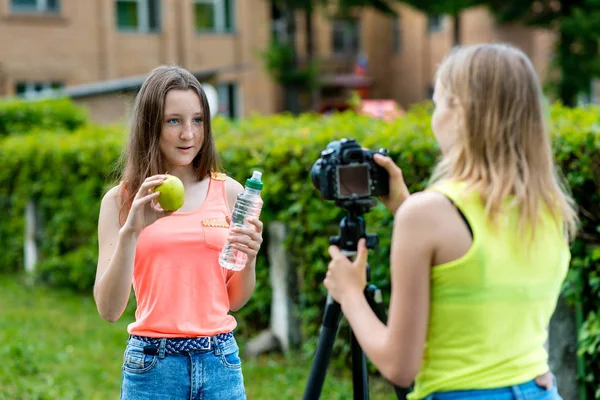 Voor meisje schoolmeisjes, zomer in de natuur. Registreert een video over gezond eten. In zijn handen houdt een groene appel en een fles water. Record Videolessen voor Internet. Gebruik camera met statief. — Stockfoto