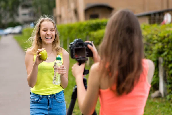 Twee meisjes zijn vriendinnen. Zomer in de natuur. Schrijft video camera. In haar handen houdt fles water en groene appel. Het concept video blog over gezonde voeding, emoties geluk is plezier. — Stockfoto