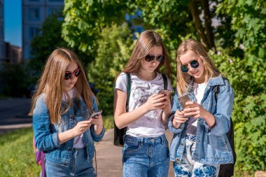 Üç kız kız öğrenci genç, kot elbise ve güneş gözlüğü takıyor. Cep telefonları tutarak ellerinde. Sosyal ağlar, çocuklarda kavramı yazıyor ve Internet iletileri okur.