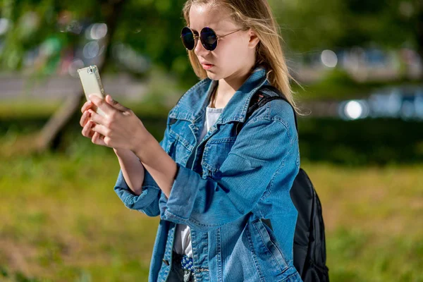 Gadis sekolahan di musim panas di taman. Di tangannya memegang smartphone. Dalam jaket denim di belakang ransel, kacamata hitam. Membaca pesan di jejaring sosial . — Stok Foto