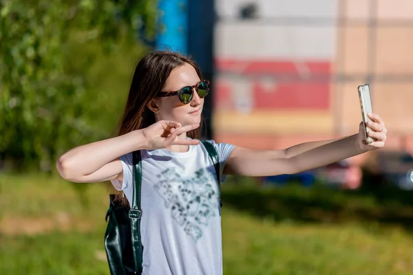 Školačka dospívající dívka, v létě v parku venku. Za batoh. V rukou drží smartphone, pořídí fotografii na telefonu. Šťastně se usmívá. — Stock fotografie