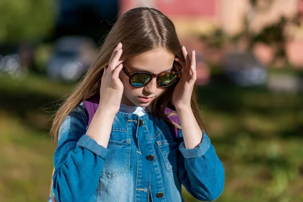 Bambina studentessa, adolescente estate all'aperto. Occhiali da sole e giacca di jeans. Corregge capelli con mani . — Foto Stock