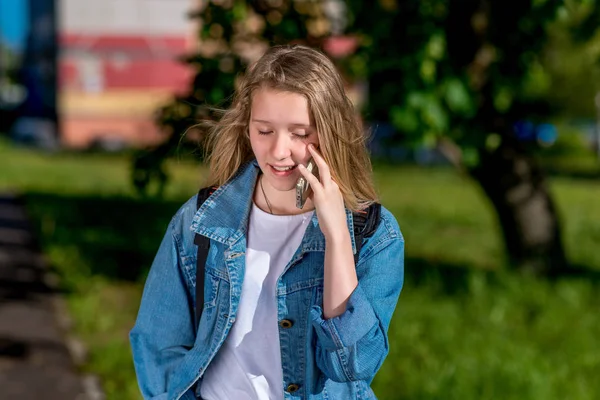 Niña colegiala adolescente, y en verano en la ciudad. Habla en el smartphone, con una chaqueta de mezclilla. Emocionalmente cierra los ojos . — Foto de Stock