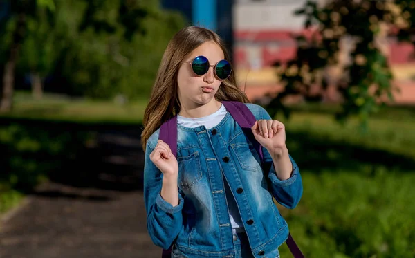 Gadis sekolah cantik remaja, di musim panas di taman di alam, mengenakan kacamata hitam di belakang ransel dan mengenakan jaket denim. Konsep lelucon dan menyenangkan, imitasi. Emosi main-main, bersenang-senang, bermain . — Stok Foto