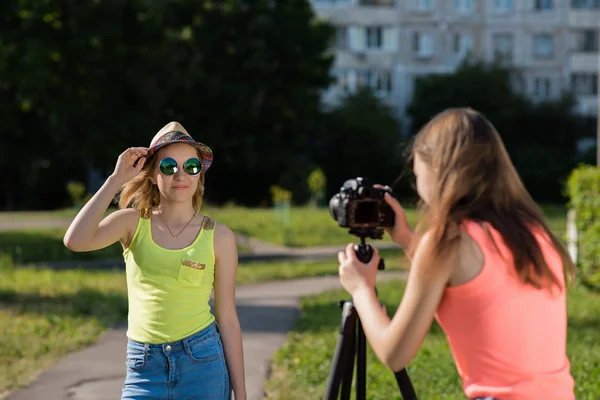 Δύο φίλες μαθήτριες το καλοκαίρι στο πάρκο ρεκόρ βίντεο στη φωτογραφική μηχανή. Αυτή είναι ντυμένη στα φωτεινά ρούχα με ένα καπέλο και γυαλιά ηλίου. Η έννοια του νέους bloggers. — Φωτογραφία Αρχείου