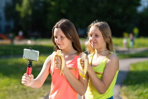 Kde jsou holčičky školačky. V létě v parku ve svých rukou drží banány. Zaznamenává video na chytrém telefonu. Šťastné mladé bloggerů se usmívají. — Stock fotografie