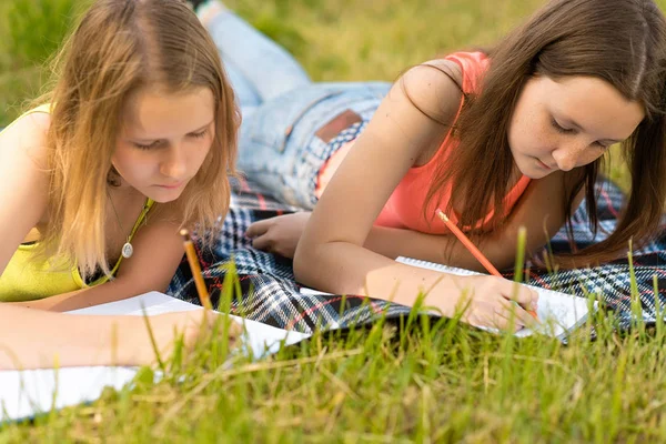 2 alunas no verão no parque estão deitadas em uma planície, fazendo trabalhos de casa em um caderno. Conceito de descanso após a escola na natureza. Emoção de concentração de estudo e conhecimento . — Fotografia de Stock