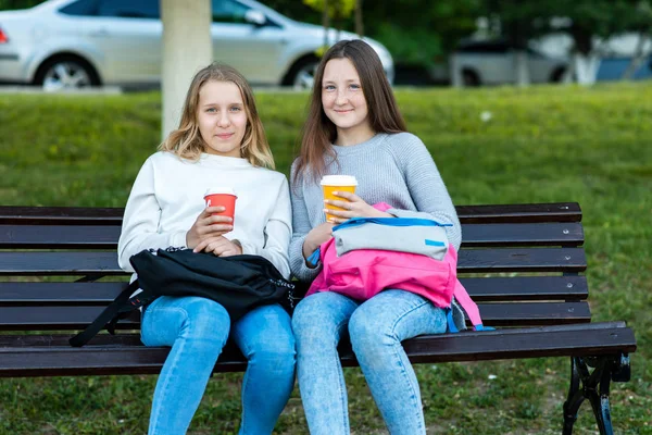 Δύο φίλες μαθήτρια κορίτσι. Το καλοκαίρι στο πάρκο κάθεται σε ένα παγκάκι. Στα χέρια του κρατά σακίδια, γυαλιά με ζεστό καφέ ή τσάι. Διάλειμμα για γεύμα μετά από το Ινστιτούτο. Χαμόγελα ευτυχώς. — Φωτογραφία Αρχείου