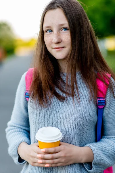 Adolescente colegial. No verão no parque. Em suas mãos segura um copo de café ou chá atrás de sua mochila. Lindos olhos azuis e cabelos longos. Close-up de um sorriso . — Fotografia de Stock