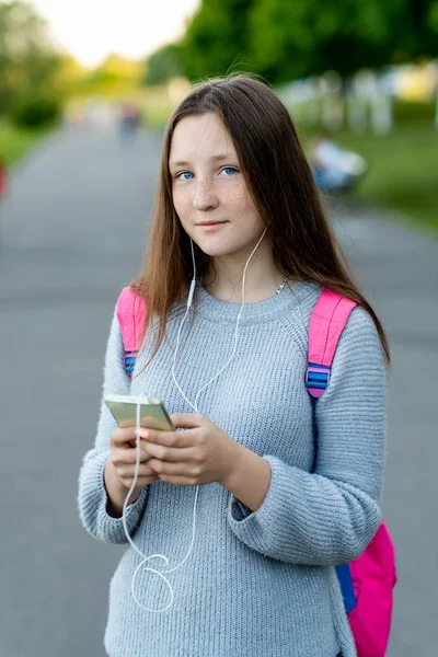 屋外の公園で美しい十代の少女の夏。手でバックパックの後ろにヘッドフォンで音楽を聴くスマート フォンを保持します。美しい青い目と長い髪. — ストック写真