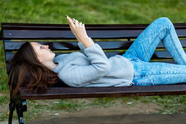 Девочка-брюнетка летом в парке лежит на скамейке. В руках у смартфона читает и пишет для переписки в социальных сетях. Отдых после школы на природе . — стоковое фото