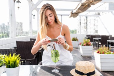 Beyaz elbiseli bir kız. Yaz parlak güneşli bir günde verandada bir kafede. Telefonda bir bardak meşrubat fotoğraf. Sosyal ağlarda sonrası. Fotoğraflar Internet için. Tabaklanmış deri uzun saçlı.