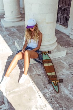 Verandada oturan güzel sarışın kız. Yakınlarda bir kaymak, longboard var. Elinde tutan bir akıllı telefon, sosyal ağlarda denk. Dersten sonra dinlenme.