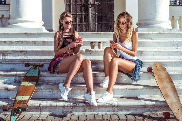 उन्हाळ्यात दोन मुली मैत्रिणी पायर्यावर विश्रांती घेतात. जवळपास स्केटबोर्ड, लाँगबोर्ड आहेत. स्मार्टफोन धारण हातात, सामाजिक नेटवर्कशी संबंधित, इंटरनेटवर संप्रेषण संकल्पना . — स्टॉक फोटो, इमेज