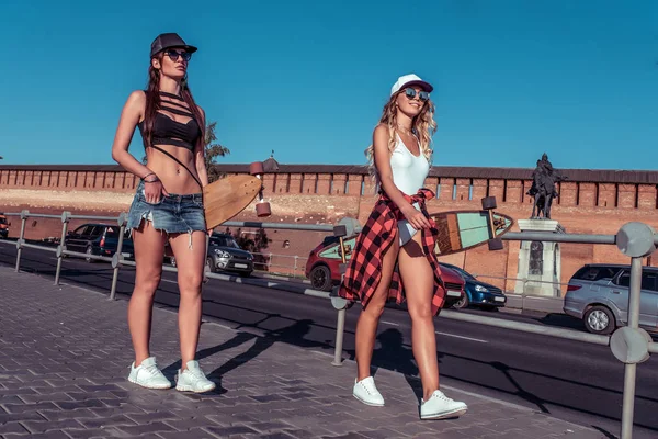 Duas amigas no verão vão à cidade. Tenho patins nas mãos. Vestida com fatos de banho leves. Verão dia ensolarado brilhante. Caminhe pela cidade. O conceito de descanso na cidade . — Fotografia de Stock