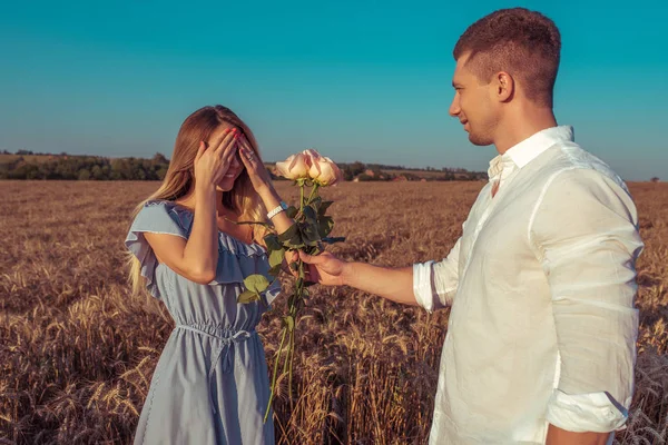 O tipo dá flores à rapariga, rosas. O verão é um dia ensolarado brilhante em um campo de trigo. O conceito de um encontro romântico. Emoções felicidade surpresa, sorriso feliz em seu rosto . — Fotografia de Stock