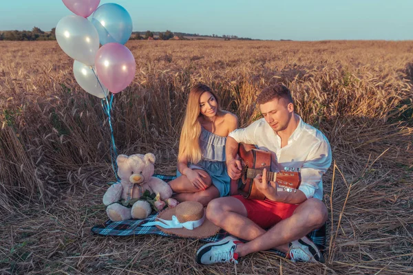 Ein junges Paar im Sommer im Weizenfeld sitzend. romantischer Abend, Erholung im Freien. das Konzept der Liebe Musik Gitarre. das Konzept von Geschenk und Urlaub. Emotionen sind Vergnügen und Zärtlichkeit. — Stockfoto