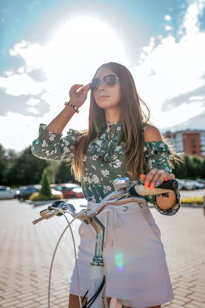 Девушка летом в городе, стоящая рядом с велосипедом, в белой юбке и зеленой блузке. Яркий солнечный день в солнечных очках . — стоковое фото