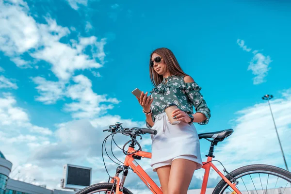 Gadis di musim panas di kota, berdiri di samping sepeda merah, dengan rok putih dan blus hijau. Dalam rukaz memegang telepon, membaca pesan di Internet. Dalam kacamata hitam . — Stok Foto