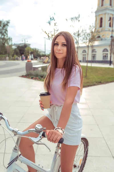 Το κορίτσι με το ποδήλατο, το καλοκαίρι στην πόλη, στα χέρια της κρατώντας ένα ποτήρι του τσαγιού. Σε ένα ροζ μπλουζάκι, όμορφο χαμόγελο. — Φωτογραφία Αρχείου