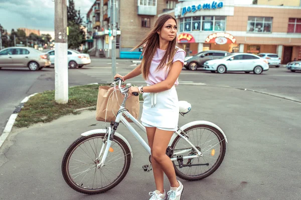 A girl stands at a crossroads during the summer in city. Pass the car at the traffic lights. With a package of products. In white skirt and pink sweater. Long beautiful hair.