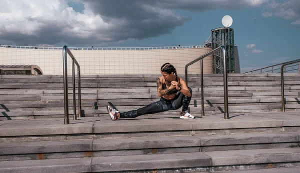 Seorang gadis dalam pakaian olahraga melakukan pemanasan sebelum jogging. Terhadap latar belakang tangga, di musim panas di kota. Gaya hidup olahraga dan sehat. Tato di tubuhnya. . — Stok Foto