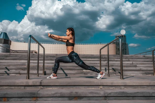 A menina com tatuagens no sportswear faz um aquecimento antes da ginástica. A fazer ioga. Contra o fundo das escadas, no verão na cidade. Esportes e estilo de vida saudável . — Fotografia de Stock