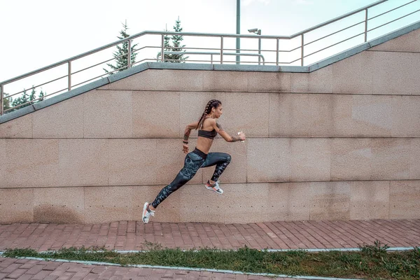 Girl athlete runs in a jump. In city on a summer day. In his hand, the player listens to music on headphones. Sports and healthy lifestyle. Free space for text.