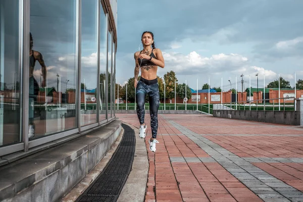 A atleta corre num salto. Na cidade em um dia de verão. Em sua mão, o jogador ouve música em fones de ouvido. Tatuagens na pele bronzeada. Esportes e estilo de vida saudável. Espaço livre para texto . — Fotografia de Stock