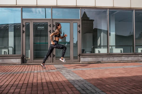 Girl athlete runs in a jump. Outside the city in morning. In his hand, a smartphone listens to music on headphones. Tattoos on tanned skin. Sports and healthy lifestyle. Free space for text.