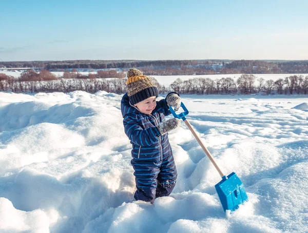 Маленький мальчик 3 лет, зимой в парке, играет с лопатой, копает снег. Счастливого светлого солнечного дня . — стоковое фото