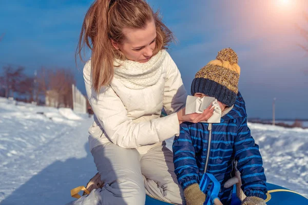 A young mother wipes her nose with a napkin to her little son 3 years old. In winter on nature in the park. Recreation and tubing. Blows his nose in a white napkin. Snot nose in winter at the boy.