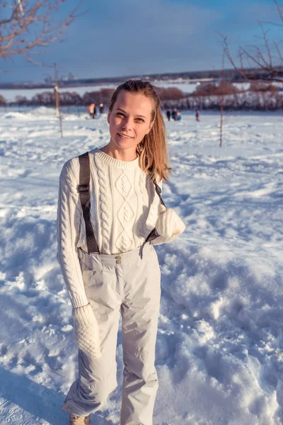Krásná mladá dívka v bílé kombinéze. V zimě na čerstvém vzduchu. Pózuje v bílém svetru a rukavice. Šťastný úsměv, odpočinek v letovisku v zasněžené počasí. — Stock fotografie