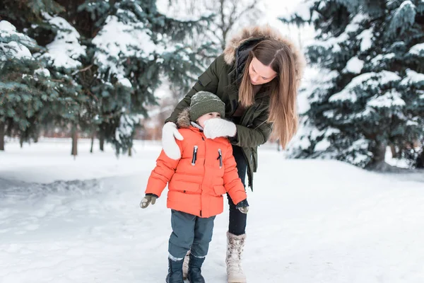 3-5岁的小孩, 一个男孩在冬天穿着温暖的夹克和帽子。冬天, 在雪中的绿色圣诞树的背景下。妈妈摇掉了宝贝的雪。他擦鼻子, 从鼻涕. — 图库照片